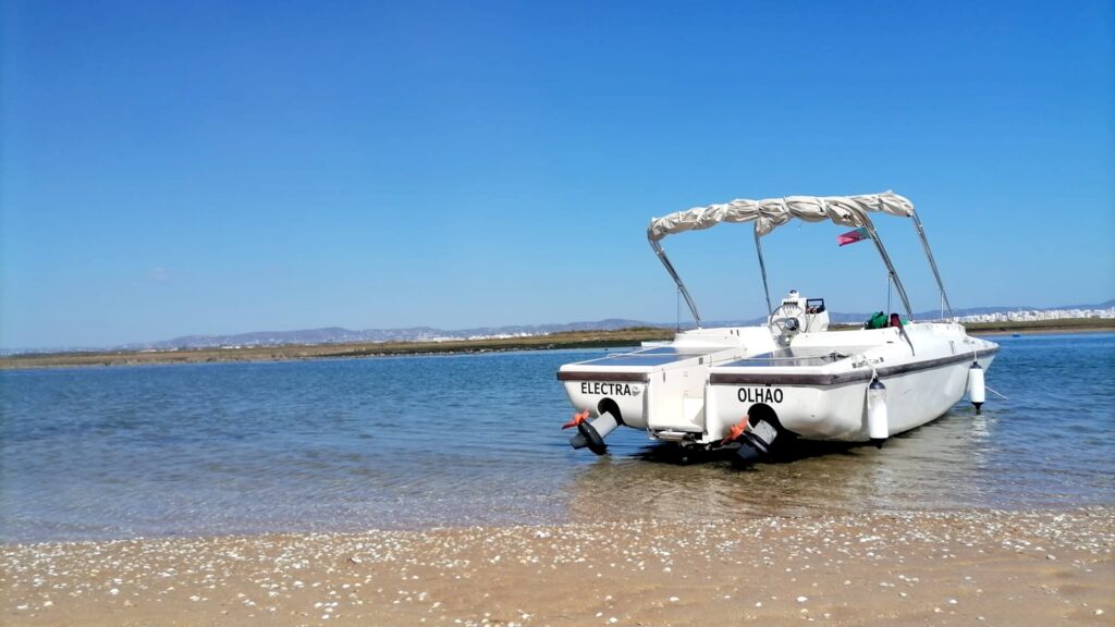 Un bateau blanc à énergie solaire avec un auvent se trouve entre le rivage et l'eau. Le ciel est bleu et le bateau a deux panneaux solaires à l'arrière. 