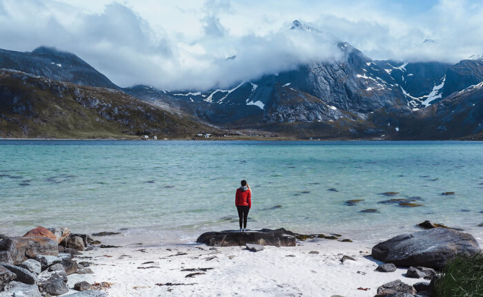 Imagem de um viajante parado junto às águas costeiras enevoadas nas Ilhas Lofoten, Noruega – ao fundo, altas montanhas avultam