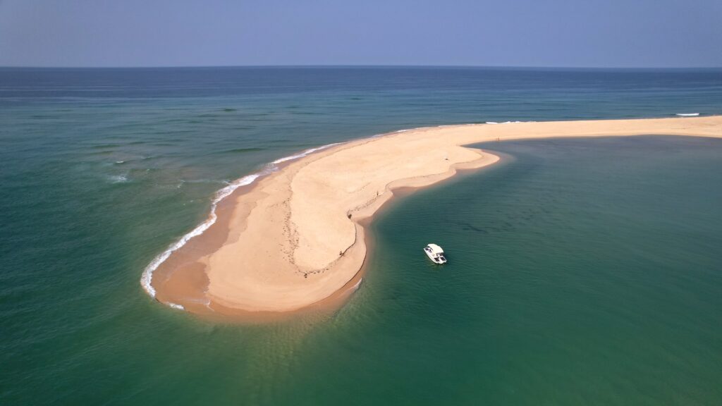 Aerial view of a Ria Formosa sandy peninsula curving into the blue ocean, with a solar-powered boat and distant figures on the beach.